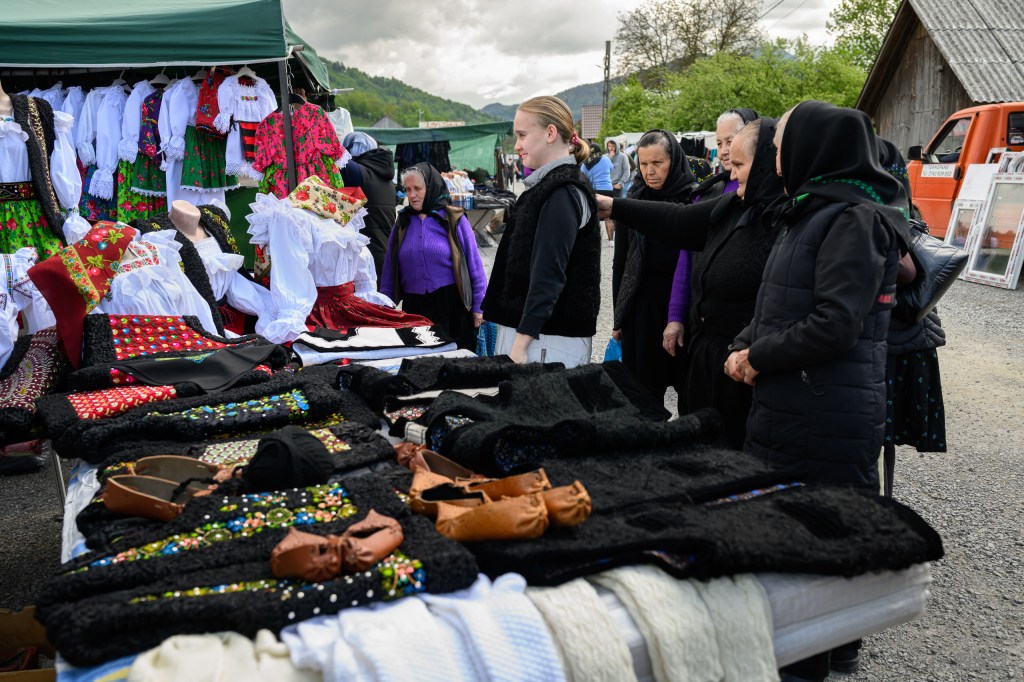 A traditional market scene with women examining colorful garments and accessories, such as dresses and vests, displayed on a table. The background features market stalls and hills.