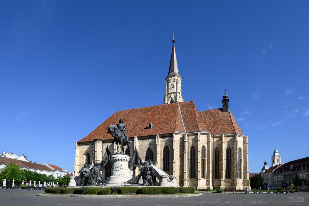 Historic church with a tall spire and an ornate roof, surrounded by statues and greenery on a clear blue sky.