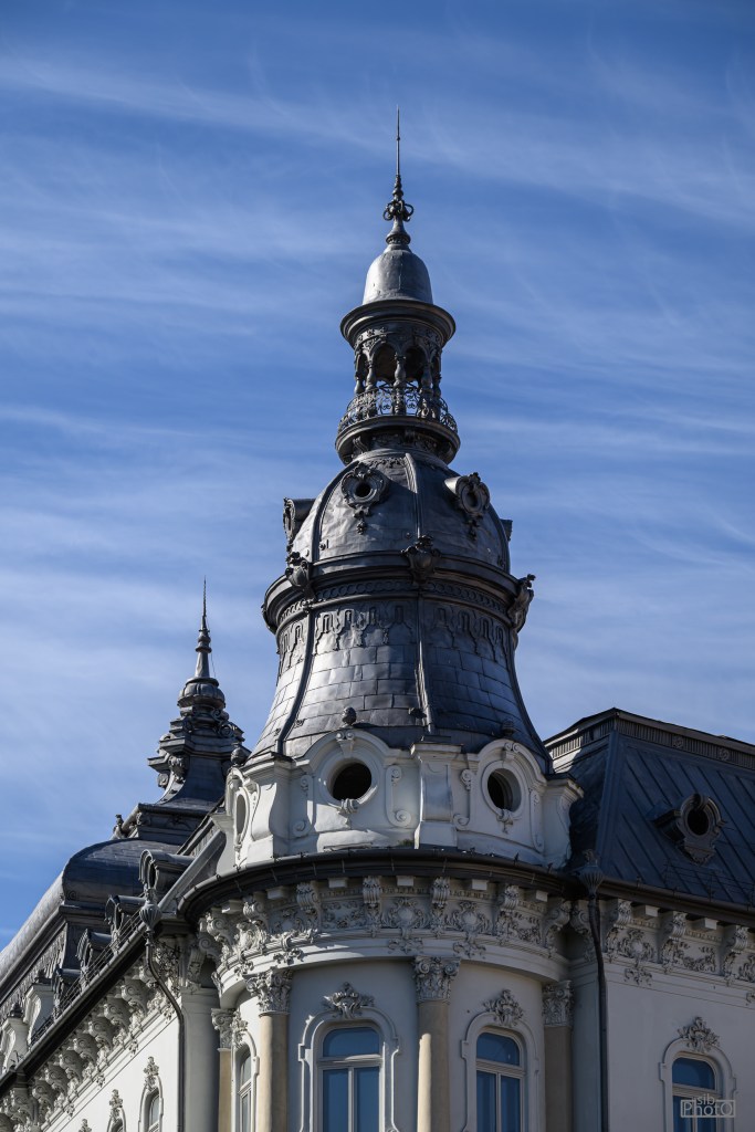 Close-up of a decorative building rooftop with a prominent, ornate spire against a blue sky.