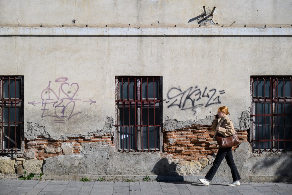 A person with red hair walks past a graffiti-covered wall featuring metal barred windows and exposed brick.