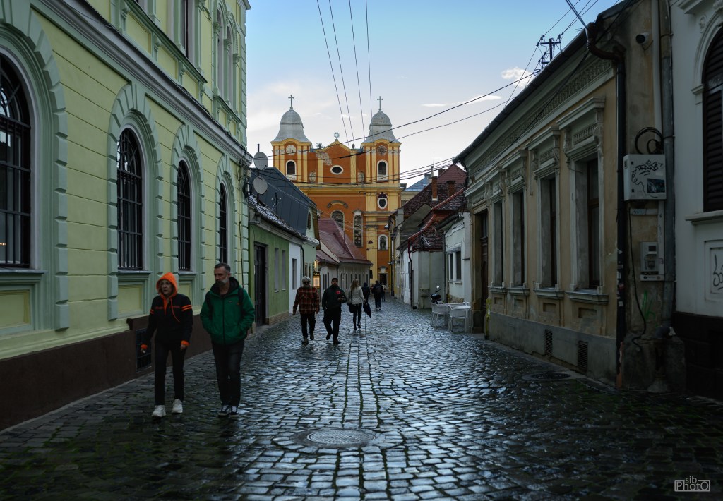 Narrow cobblestone street with people walking, featuring colorful buildings and a church in the background under a cloudy sky.