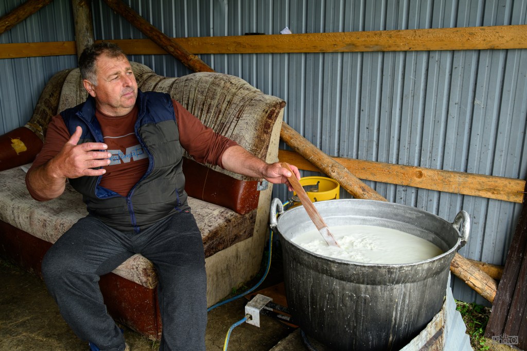 A man sitting on a couch stirring a large pot of milk with a wooden spoon in a rustic indoor setting.