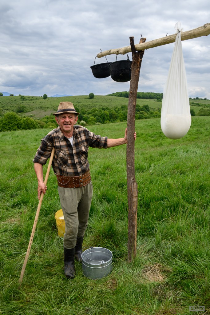 An older man wearing a hat and plaid shirt stands in a grassy field next to a wooden post supporting two hanging black buckets and a large white bag. He holds a stick and smiles at the camera, with a green landscape and cloudy sky in the background.