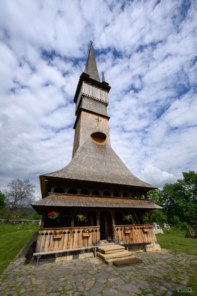 A traditional wooden church with a tall spire, featuring intricate architecture and flower planters, set against a cloudy sky.