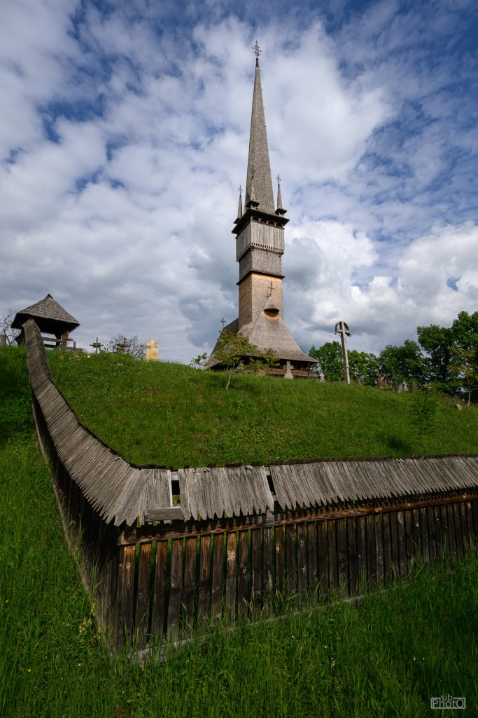 A tall wooden church tower with a pointed roof stands on a grassy hill, surrounded by a low wooden fence under a partly cloudy sky.