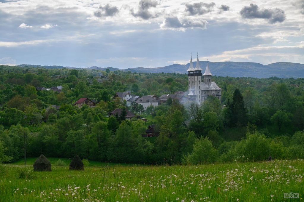 A panoramic view of a lush green landscape with a village in the foreground and a church with tall spires in the background, under a cloudy sky.