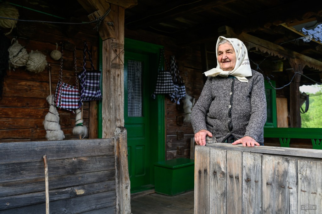 An elderly woman stands at the entrance of a rustic wooden house, smiling and wearing a headscarf. The house is adorned with traditional decorations, including handmade items hanging on the walls.