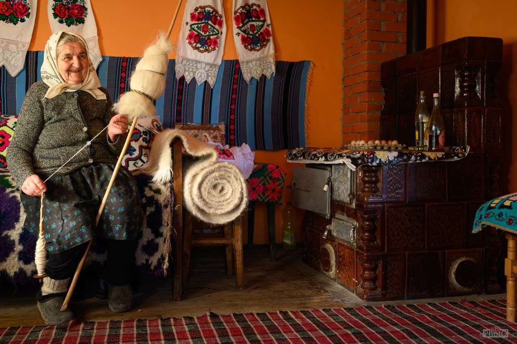 An elderly woman sitting on a colorful patterned couch, spinning wool with a spindle in her hand. She is wearing a traditional outfit with a headscarf. Beside her, there are rolls of wool, and in the background, there is a rustic stove and decorative textiles hanging on the wall.