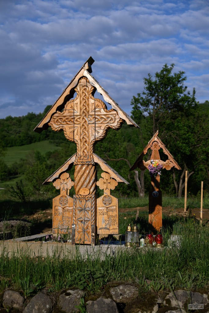 Carved wooden cross and gravestones in a cemetery setting, surrounded by greenery and under a blue sky with clouds.