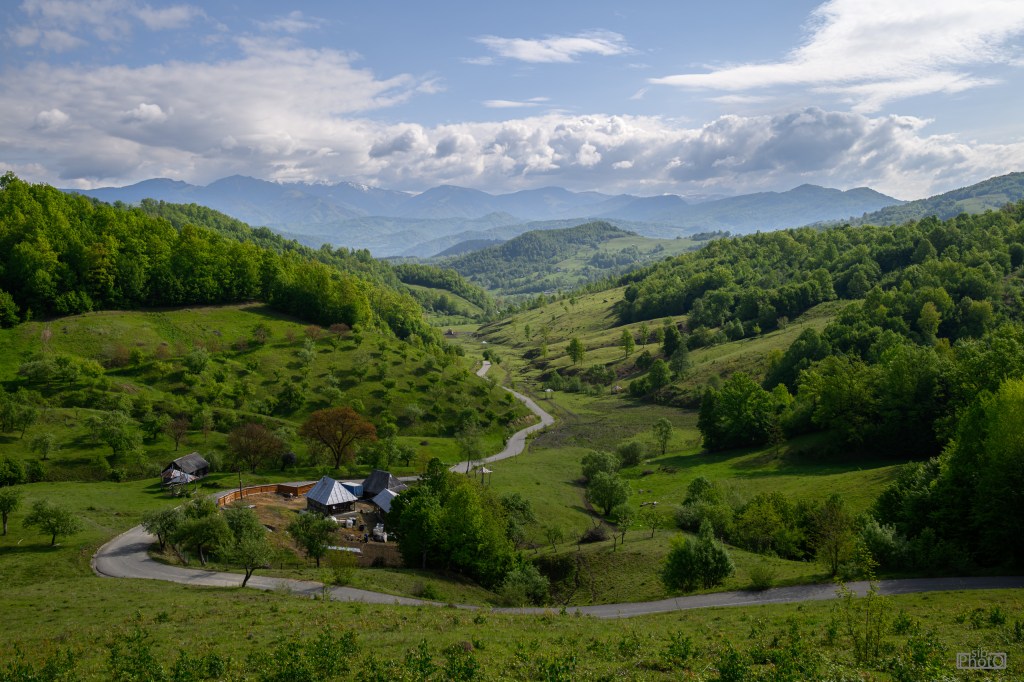 A scenic view of rolling green hills and a winding road, with a small cluster of buildings at the base and mountains in the background under a partly cloudy sky.