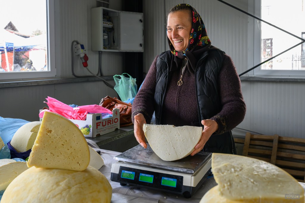A smiling woman wearing a headscarf is standing at a market stall, holding a large piece of cheese on a scale while various cheeses are displayed around her.