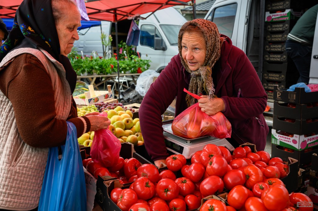 A market scene featuring two women engaging in a transaction while surrounded by fresh produce, including a variety of tomatoes and apples.