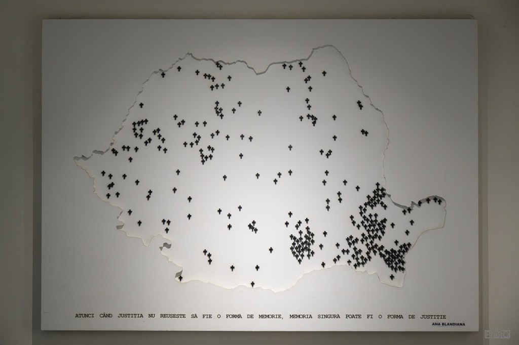 A white map of Romania with numerous black crosses scattered across it, symbolizing various locations. The bottom features a Romanian quote about justice and memory.
