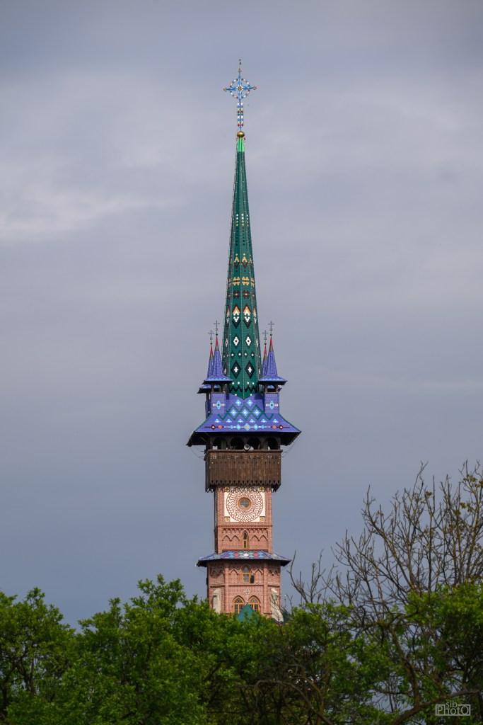 Close-up view of a colorful ornate steeple with a cross on top, surrounded by green trees under a cloudy sky.