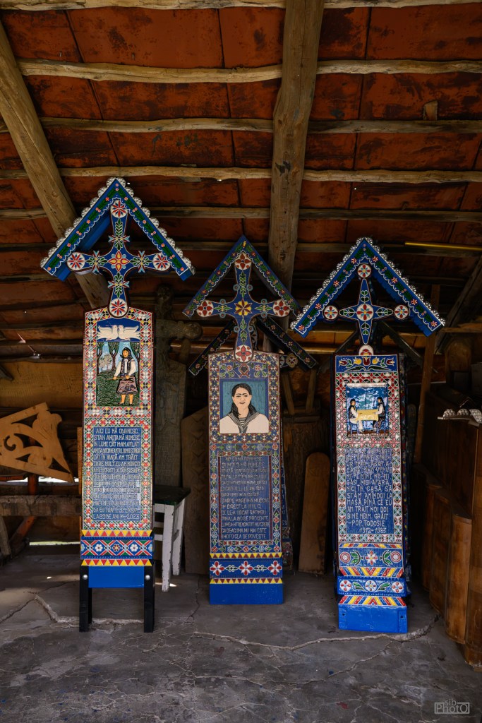 Three intricately carved wooden memorial crosses with colorful decorations, featuring portraits and inscriptions, set against a rustic wooden backdrop.