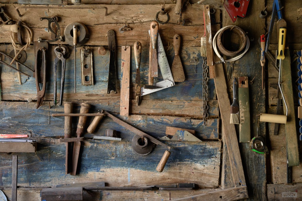A collection of various hand tools arranged on a rustic wooden wall, featuring pliers, saws, hammers, and measuring devices.