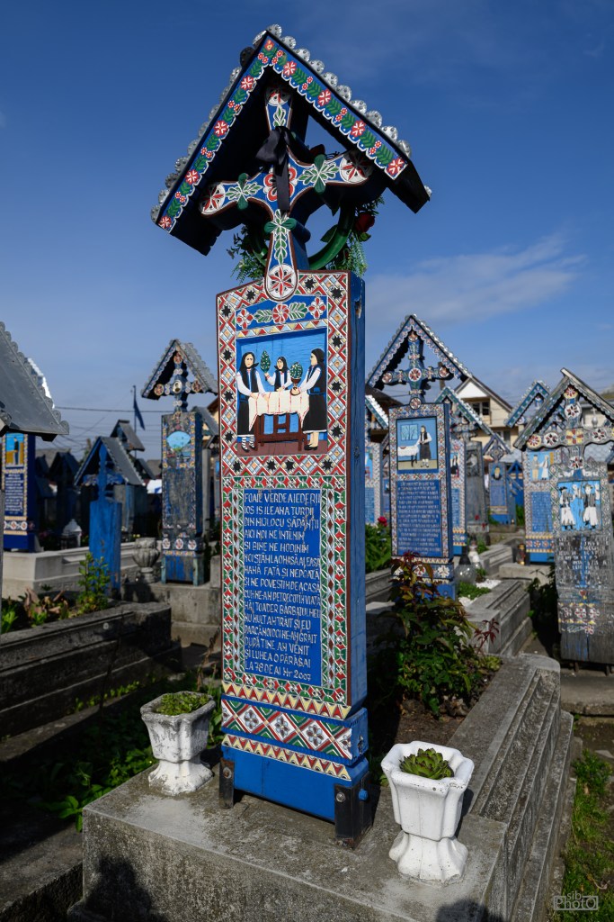 A colorful wooden tombstone adorned with intricate patterns and a painted scene, standing in a cemetery filled with similar markers. The tombstone features a cross at the top and is surrounded by flower pots.