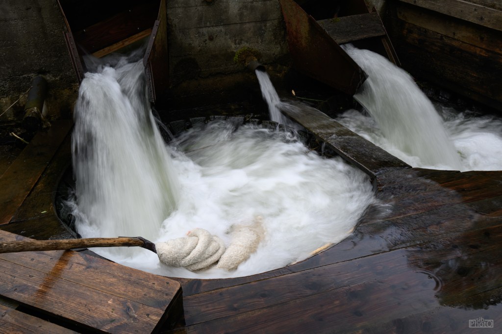 A water wheel mechanism with flowing water creating white rapids around its circular base, surrounded by wooden structures.