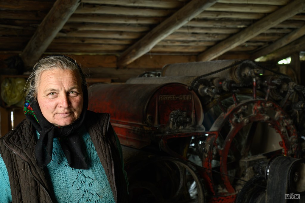 An older woman wearing a scarf and sweater stands in a rustic shed with a vintage farming machine in the background.