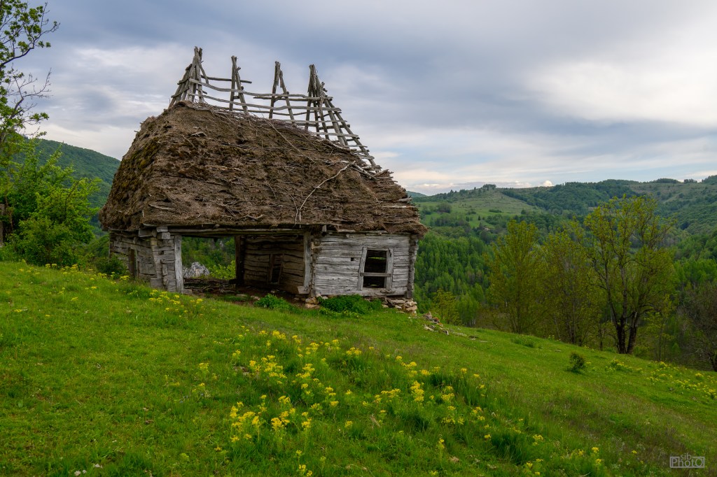 An old, partially collapsed wooden house with a thatched roof, surrounded by lush green grass and trees in a hilly landscape under a cloudy sky.