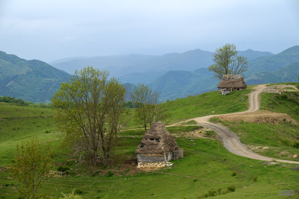 Scenic view of a rural landscape featuring two traditional huts on a hill, with green fields and distant mountains under a cloudy sky.