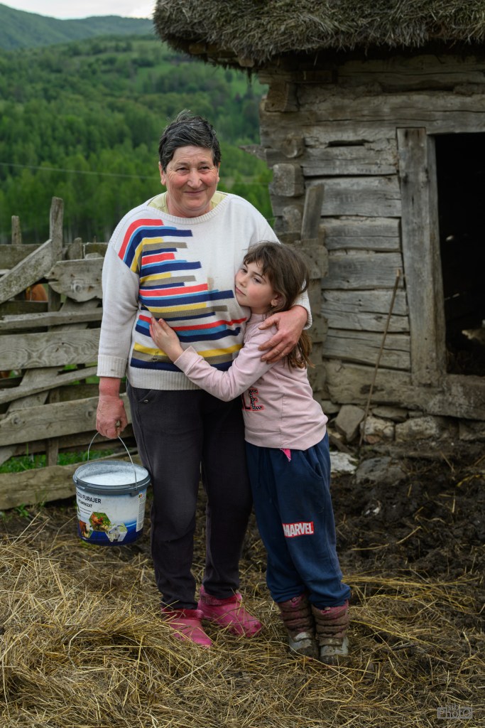 An older woman wearing a colorful sweater poses with a young girl in a rural setting, standing in front of a wooden barn. The woman holds a bucket and the girl hugs her side.