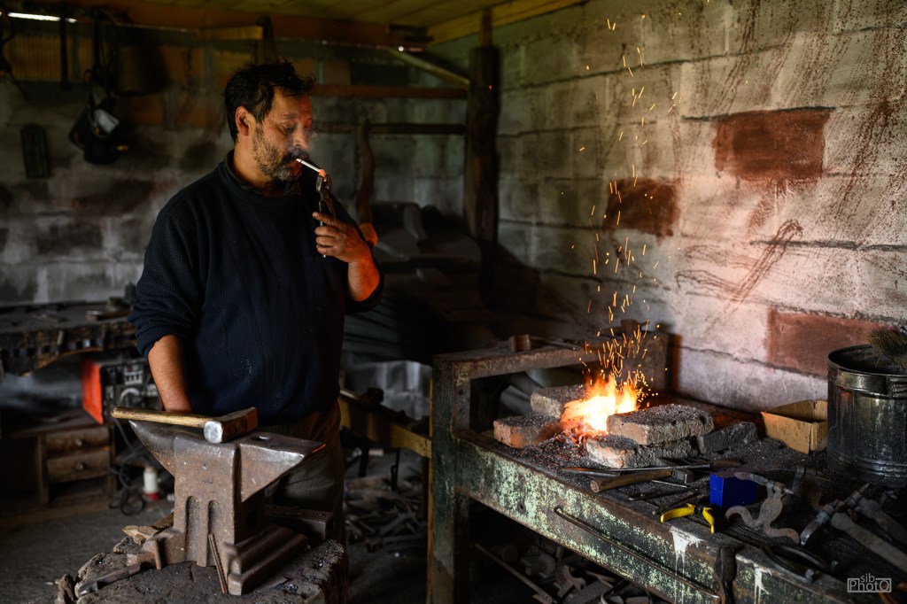 A blacksmith working in a workshop, using a blowtorch while sparks fly from a small fire on a stone surface. Tools and an anvil are visible on the workbench.