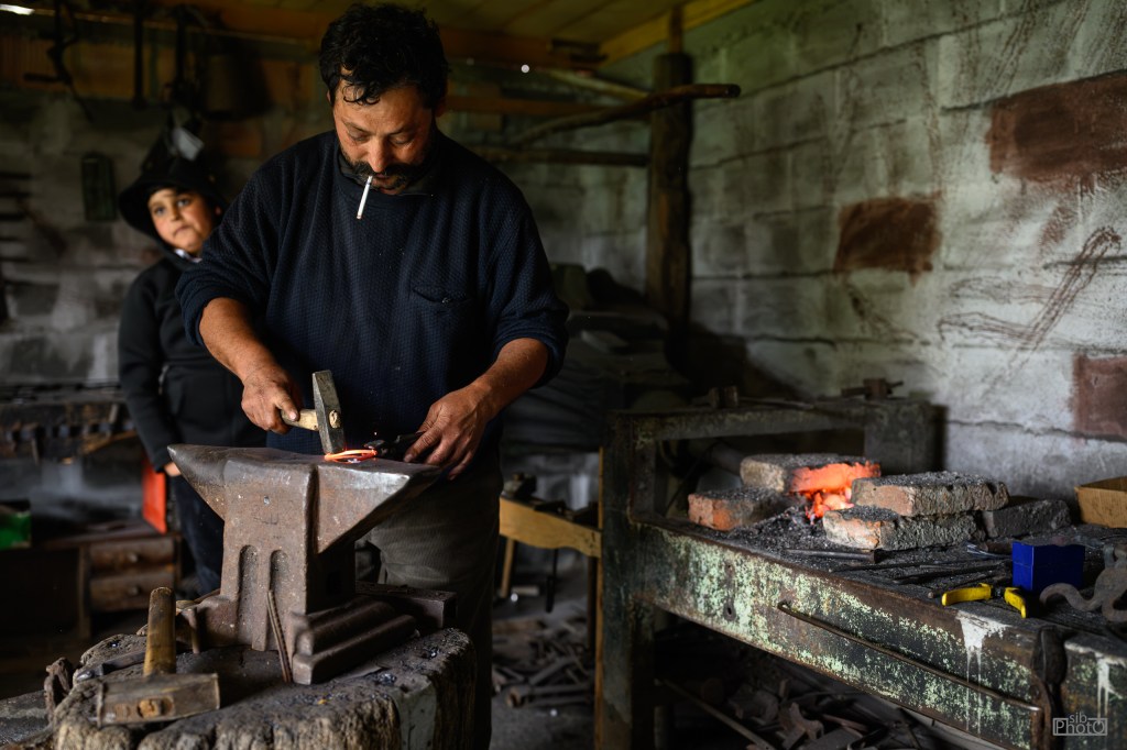 A blacksmith shaping hot metal on an anvil in a workshop, with a woman watching in the background.