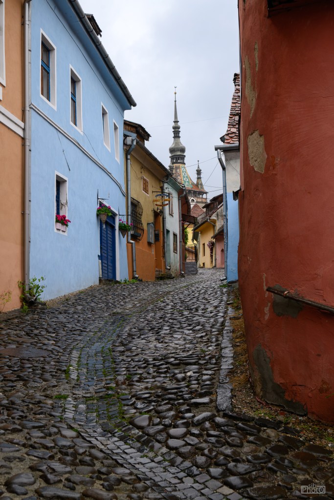A narrow cobblestone alley lined with colorful buildings, featuring a church spire in the background under a cloudy sky.