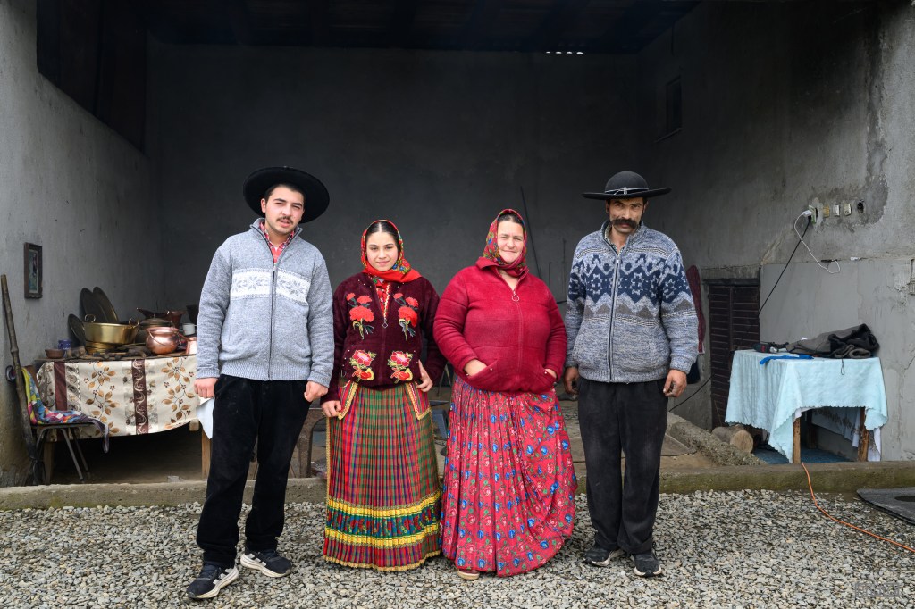 A group of four individuals dressed in traditional clothing standing outside a rustic building. The men wear sweaters and hats, while the women are in brightly colored dresses and shawls. Background features cooking utensils and an informal outdoor setting.