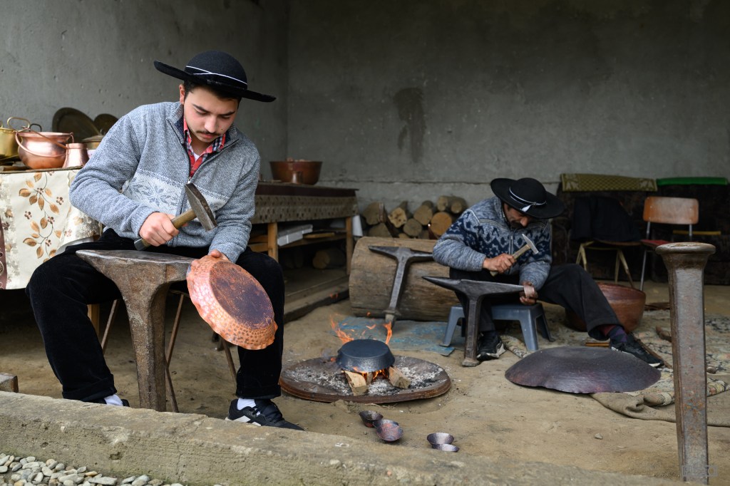Two artisans working on metal crafting in a rustic workshop. One man is hammering a copper dish on an anvil, while the other is shaping a metal object, with tools and materials visible around them.