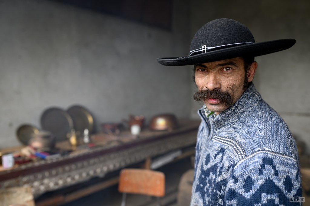 A man with a distinctive mustache wears a traditional black hat and a patterned sweater, looking towards the camera with a serious expression in a rustic setting.