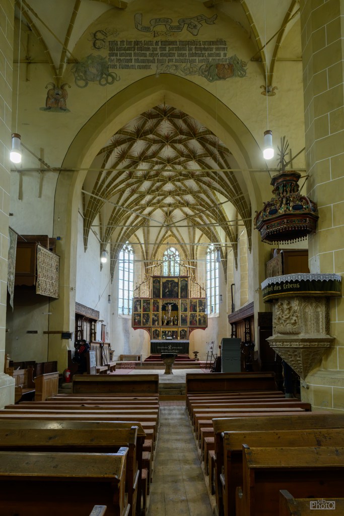 Interior of a historic church featuring wooden pews, an altar with a tall painted backdrop, and intricately designed arches and ceiling, illuminated by natural light from large windows.
