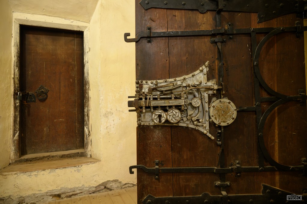 Close-up view of a vintage wooden door with intricate metalwork and locking mechanism, alongside a smaller wooden door in a dimly lit room.