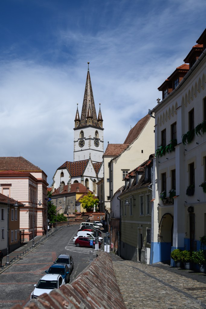 A picturesque street view with a cobblestone road lined with parked cars, and buildings featuring historical architecture. In the background, a tall church with a distinct spire is visible against a blue sky.
