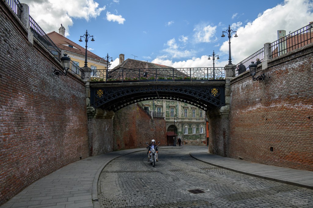 A narrow cobblestone street leading under a decorative arch bridge, flanked by brick walls and historic buildings under a partly cloudy sky.