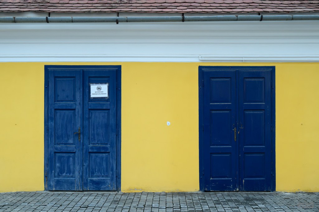 Two blue wooden doors against a yellow wall, with a notice posted on the left door.