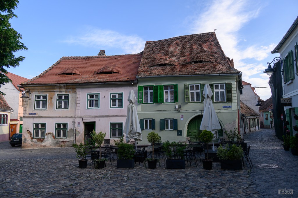 Historic buildings with colorful facades and shutters, featuring outdoor seating and umbrellas in a cobblestone square.