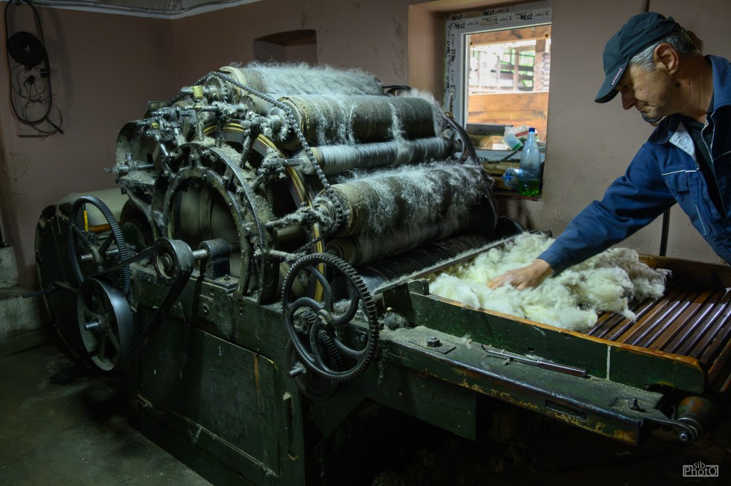 A man operating a wool carding machine, with wool fluff being processed on the machine's surface in a dimly lit workshop.