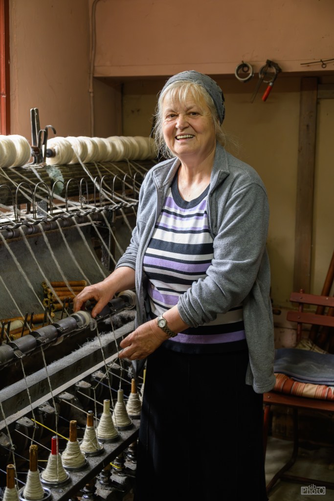 An elderly woman smiling while working at a traditional weaving machine, with spools of thread in the background.