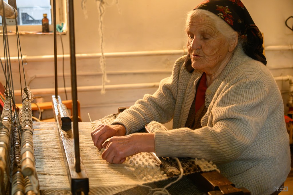 An elderly woman weaving fabric on a traditional loom, focused on her work in a well-lit room.
