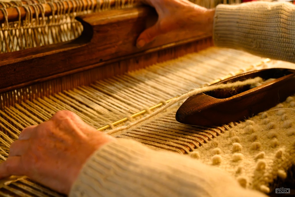 Close-up of hands weaving on a loom, with a wooden shuttle and threads visible.