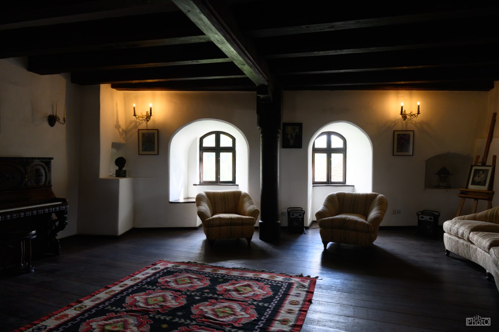 A cozy interior room featuring two striped armchairs, a patterned rug, and wooden beams. There are decorative wall sconces and two arched windows allowing natural light, with a piano and easel in the background.