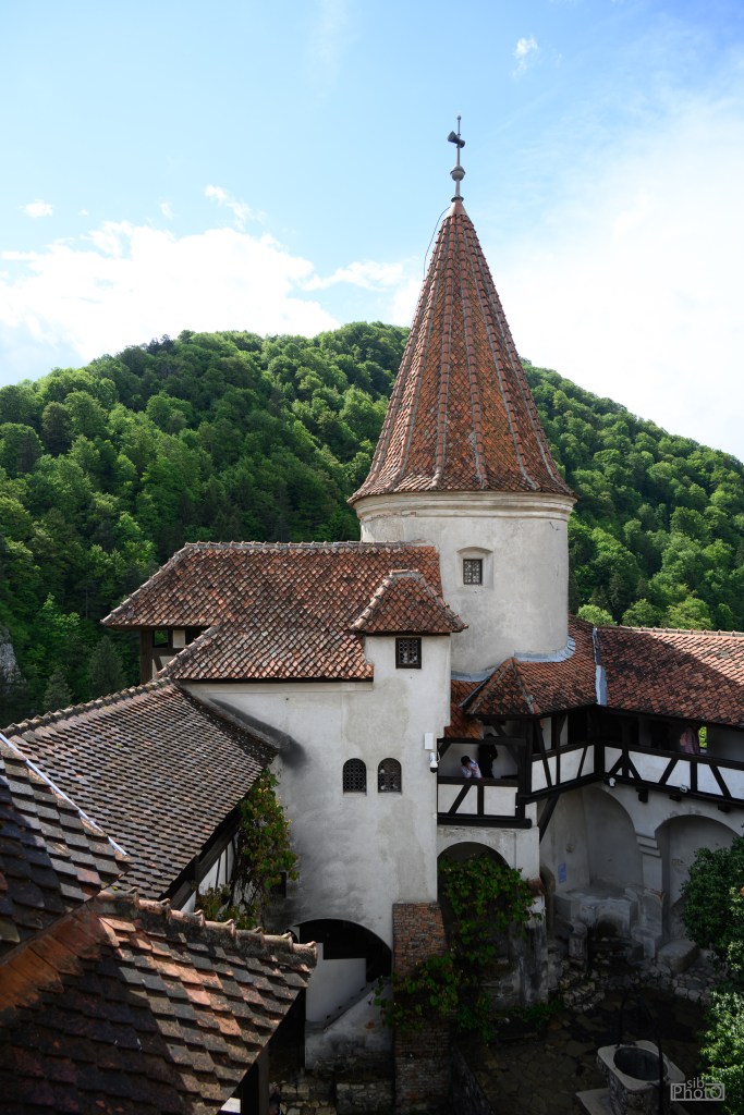 A historical building with a pointed tower and red tiled roofs, nestled among lush green hills under a blue sky.