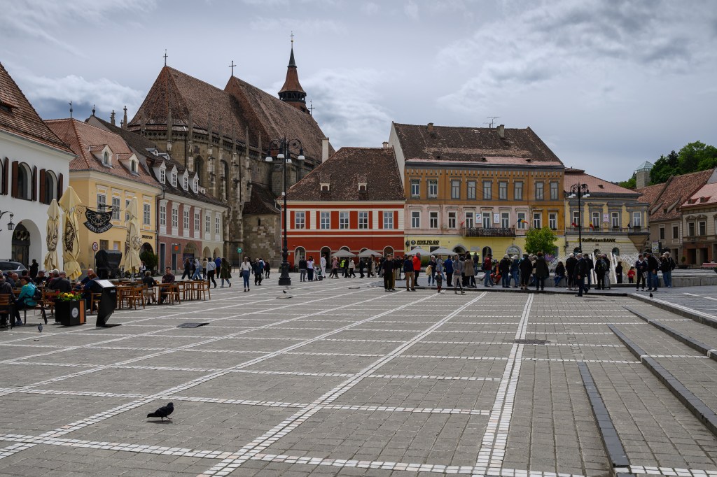 A view of a bustling public square featuring people walking, sitting at outdoor tables, and various colorful buildings, including historical architecture and shops, under a partly cloudy sky.