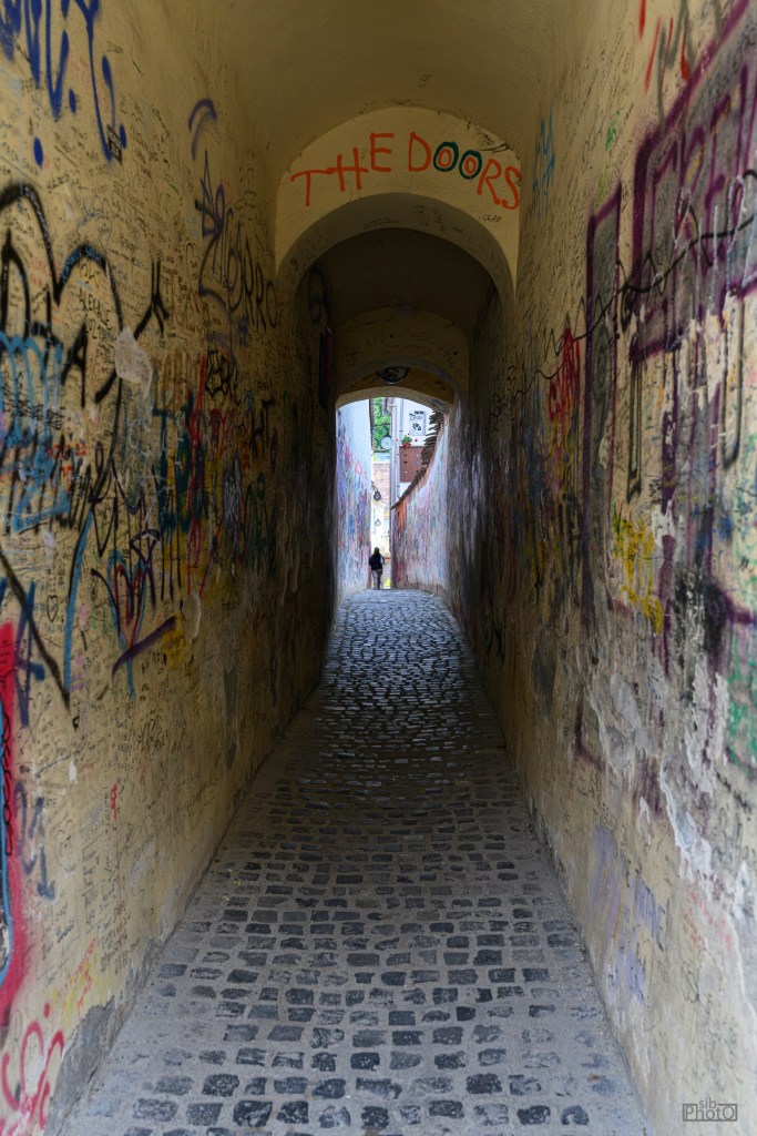 Narrow alleyway with cobblestone pavement, lined with colorful graffiti on the walls, leading to a light at the end.