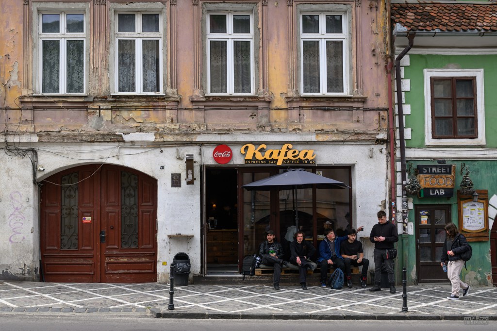 A group of young people sitting outside a coffee shop named 'Kafea,' with a faded building facade and a street sign for 'Street Food Lab' visible nearby.