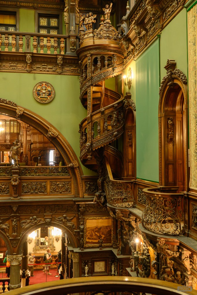 Interior view of an ornate staircase in a historical building, featuring intricate wooden carvings, green walls, and decorative sculptures.