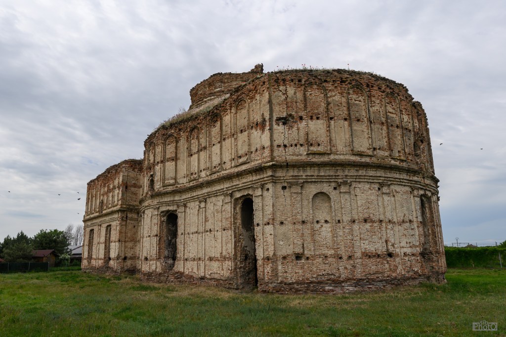 Side view of a crumbling historical brick structure with arches, set against a cloudy sky and surrounded by grassy fields.