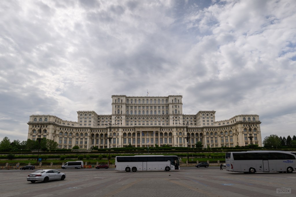 A large historical building with a grand architectural design, featuring multiple floors, columns, and balconies, set against a cloudy sky. Several buses and cars are parked in the foreground, surrounded by greenery.